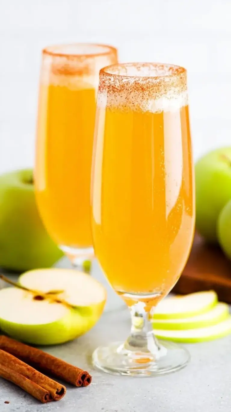 Four champagne flutes filled with golden apple cider mimosa and topped with apple slices and cinnamon sticks
